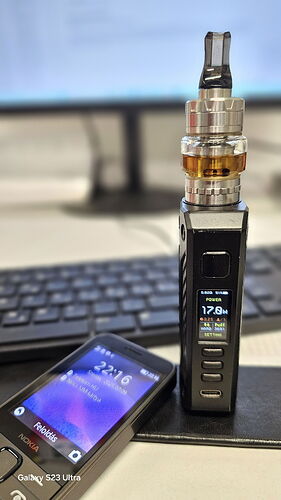 Egy asztalon egy zárt laptopon áll egy elektronikus cigaretta, mellette pedig egy mobiltelefon látható, háttérben egy homályos számítógép-képernyő és billentyűzet. (Feliratozva AI-val)