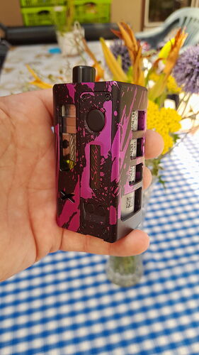 Egy kéz tart egy fekete és lila mintás elektromos cigarettát, háttérben egy kék-fehér kockás terítő és virágok láthatók. (Feliratozva AI-val)