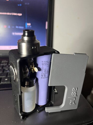 A nyitott oldallapú fekete vape mod belső részeit mutatja, beleértve egy lila akkumulátort és egy folyadéktartályt, egy laptop előtt. (Feliratozva AI-val)