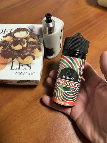 A képen egy kézben tartott Bombo Bar Juice e-liquid üveg látható, mellette egy vape eszköz és egy sütemény doboz. (Feliratozva AI-val)