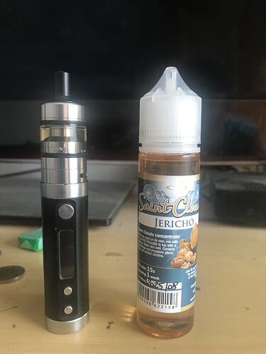 A képen egy fekete és ezüst e-cigaretta látható egy üveg "Saint Cloud Jericho" márkájú e-liquiddel együtt. (Feliratozva AI-val)
