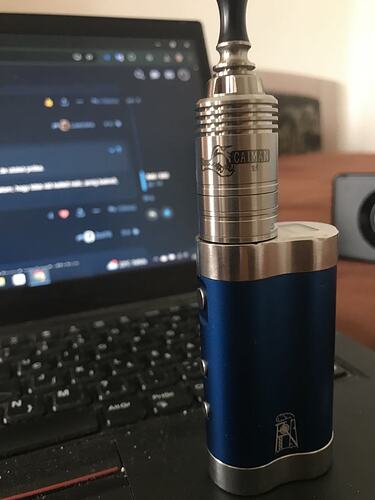 Egy kék és ezüst vape eszköz, amelyen a "CAIMAN V4" felirat olvasható, egy laptop billentyűzetén fekszik, háttérben a képernyővel. (Feliratozva AI-val)