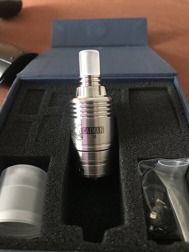 Egy ezüst színű, hengeres vaping eszköz, Caiman V4 felirattal látható egy kék, habszivacs bélésű dobozban, mellette további tartozékokkal. (Feliratozva AI-val)