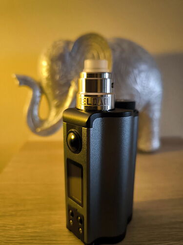 Egy fekete vape eszköz áll egy fa felületen, elmosódott ezüst elefánttal a háttérben. (Feliratozva AI-val)