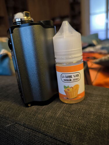 Egy sötétszürke e-cigaretta készülék és egy barack-sárgabarack ízű e-liquid palack látható egy kanapén. (Feliratozva AI-val)