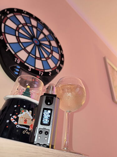 A képen egy rózsaszín falon függő dartstábla alatt egy polcon karácsonyi témájú bögre, egy vape eszköz és egy italos pohár látható. (Feliratozva AI-val)