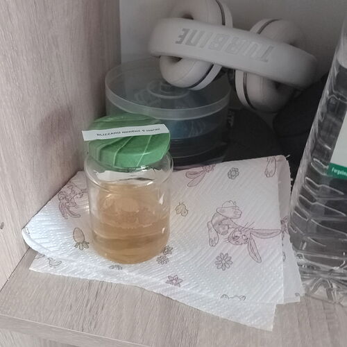 Egy üvegpohár teával egy papírtörlőn, mögötte fehér fejhallgató és egy műanyag fedeles tárolóhely található egy polcon. (Feliratozva AI-val)