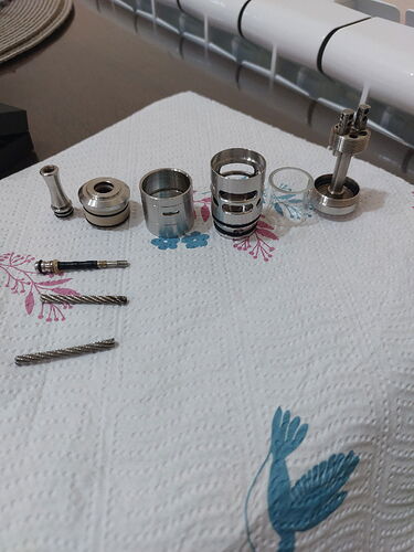 A képen különböző fém alkatrészek és csavarok vannak elhelyezve egy fehér, mintás textil felületen. (Feliratozva AI-val)