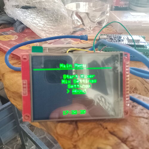 Egy közelről készült felvétel egy kis elektronikus kijelzőt mutat zöld szöveggel, amely egy menüt jelenít meg, és más elektronikus alkatrészekhez és kék vezetékekhez csatlakozik egy felületen. (Feliratozva AI-val)