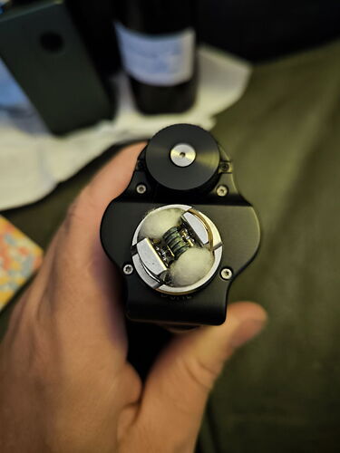 A képen egy fekete vape készülék látható, amelynek tetején kettős tekercses porlasztó található, fehér vattával. (Feliratozva AI-val)