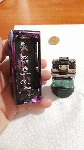 Egy kéz tart egy lila és fekete, nyitott elemtartó rekeszű vape modot, mellette egy szennyezett porlasztó látható egy fehér felületen. (Feliratozva AI-val)