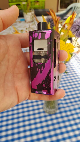 Egy kéz egy lila és fekete, mintás e-cigarettát tart egy kék-fehér kockás asztalterítő fölött, elmosódott virágokkal a háttérben. (Feliratozva AI-val)
