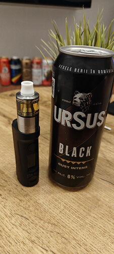 Egy fekete vape és egy Ursus Black sörösdoboz áll egy fa asztalon, a háttérben növények és további dobozok láthatók. (Feliratozva AI-val)