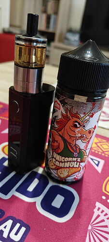 Egy fekete e-cigaretta áll egy "Unicorn's Cannoli" feliratos e-liquid flakon mellett, mindkettő egy színes, mintás alátéten fekszik. (Feliratozva AI-val)