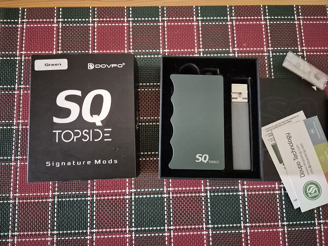 Egy Dovpo SQ Topside Signature Mods e-cigaretta készlet látható a dobozával, az eszközzel és a tartozékaival együtt egy kockás terítőn. (Feliratozva AI-val)