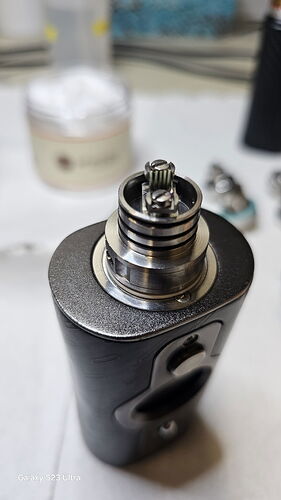 Az image egy közelkép egy elektromos vape modról, amelyen egy tekercs és vattatartó rész látható. (Feliratozva AI-val)