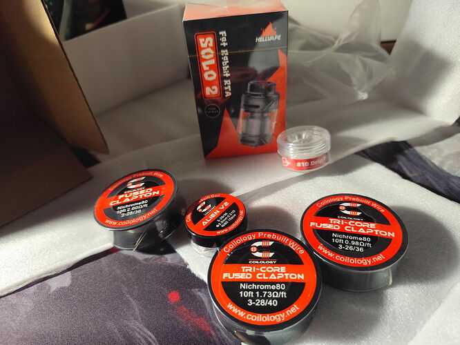 Különféle vaping kellékek, például huzaltekercsek és egy Solo 2 RTA doboz láthatók egy mintás felületen elrendezve. (Feliratozva AI-val)