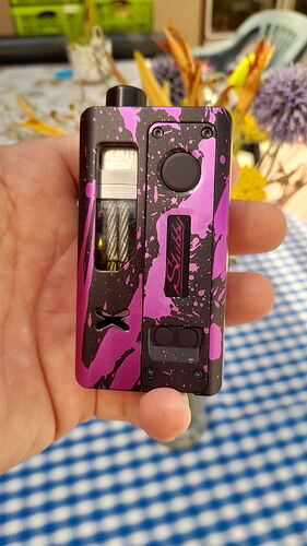 A képen egy rózsaszín és fekete fröcskölt mintás vape eszközt tartó kéz látható, a háttérben elmosódott virágokkal és egy kockás asztalterítővel. (Feliratozva AI-val)