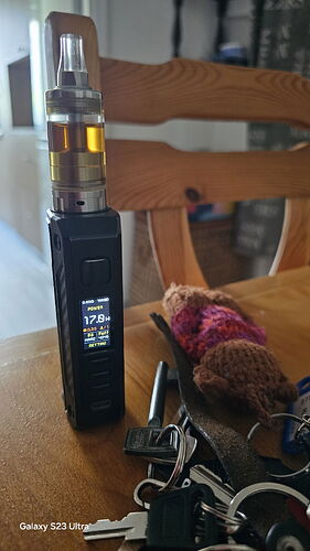 Egy elektronikus cigaretta áll az asztalon egy fa székkel a háttérben, mellette kulcsok és egy kötött holmi látható. (Feliratozva AI-val)