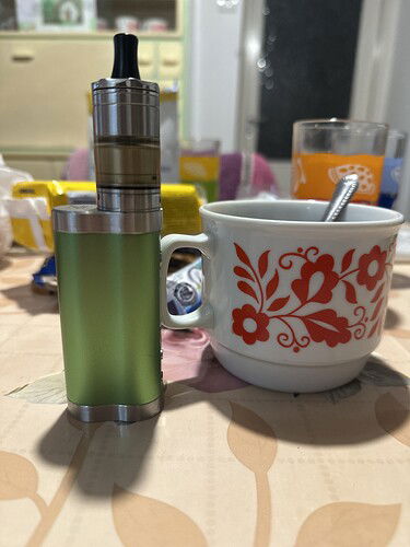 Egy zöld vape eszköz áll egy piros virágmintás fehér csésze mellett, a háttérben egy pohár narancssárga folyadékkal. (Feliratozva AI-val)