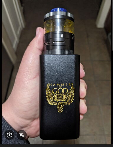 Egy személy egy fekete vape eszközt tart, arany 'HAMMER OF GOD' logóval és egy sárga folyadékkal teli átlátszó tartállyal. (Feliratozva AI-val)
