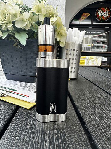 Egy vape készülék áll egy sötét színű asztalon, fehér virágokkal és evőeszközökkel a háttérben. (Feliratozva AI-val)