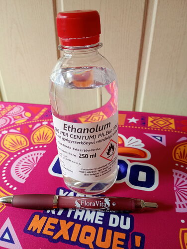 Egy 250 ml-es etanolum (etanol) oldatot tartalmazó, piros kupakos műanyag palack látható, amely egy színes, mintás felületen áll, előtte egy FloraVita toll van elhelyezve. (Feliratozva AI-val)