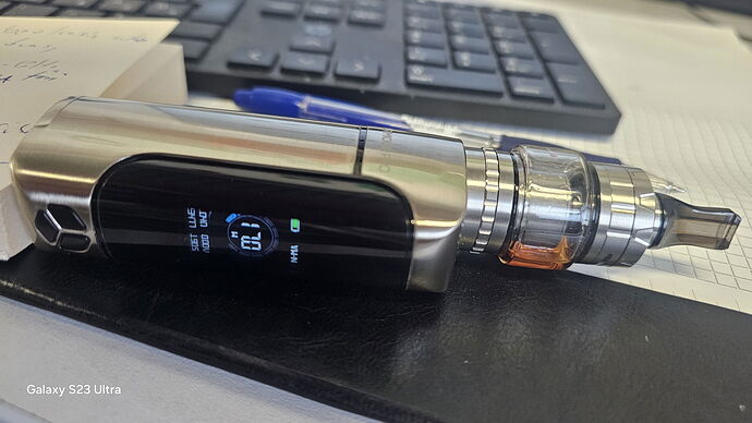 Egy ezüst színű vape eszköz fekszik egy asztalon, háttérben egy billentyűzettel és egy tollal. (Feliratozva AI-val)