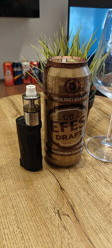 Egy fekete vape készülék és egy Efes Draft sörcan látható egy fa asztalon, egy növény, egy borospohár és további italos dobozok társaságában a háttérben. (Feliratozva AI-val)