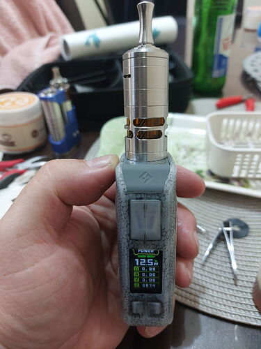 Egy kézben tartott, digitális kijelzős elektromos cigaretta (vape) látható, háttérben különféle tárgyakkal. (Feliratozva AI-val)