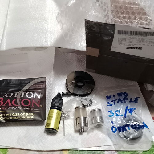 Különböző gőzölő kellékek, mint például Cotton Bacon, e-liquid és vape tartály alkatrészek, vannak elrendezve egy felületen egy csomag mellett. (Feliratozva AI-val)