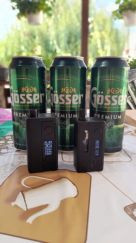 Három zöld Gösser sörösdoboz és két fekete vape eszköz látható egy asztalon, zöld növényzetes háttér előtt. (Feliratozva AI-val)