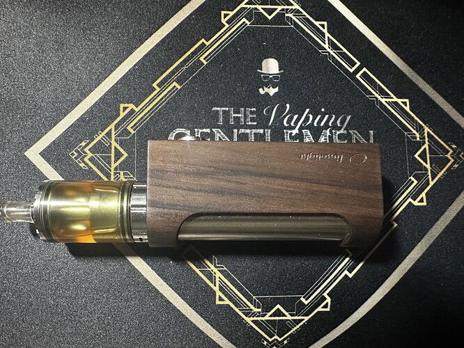 Egy fa borítású, modern kialakítású vape eszköz fekete háttéren, amelyen az angol felirat: "The Vaping Gentlemen" szerepel. (Feliratozva AI-val)