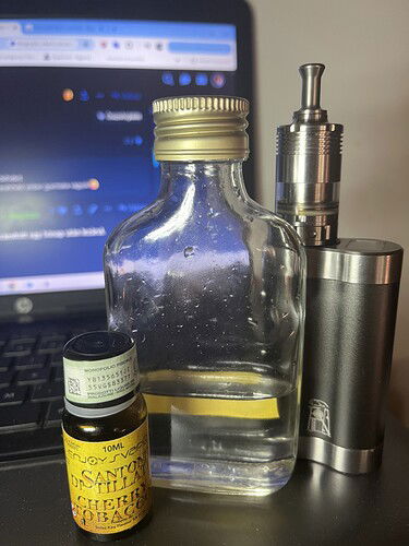 Egy asztalon egy félig telt üveg, egy elektromos cigaretta és egy illóolajos üveg áll, háttérben egy laptop képernyőjével. (Feliratozva AI-val)