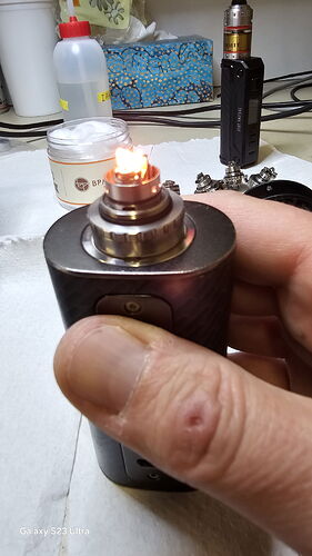 A képen egy kéz tart egy égő vape modot, háttérben különböző vape tartozékokkal és eszközökkel. (Feliratozva AI-val)